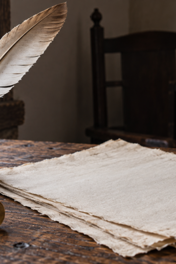 Blank handmade paper and a quill rest on a colonial oak table.
