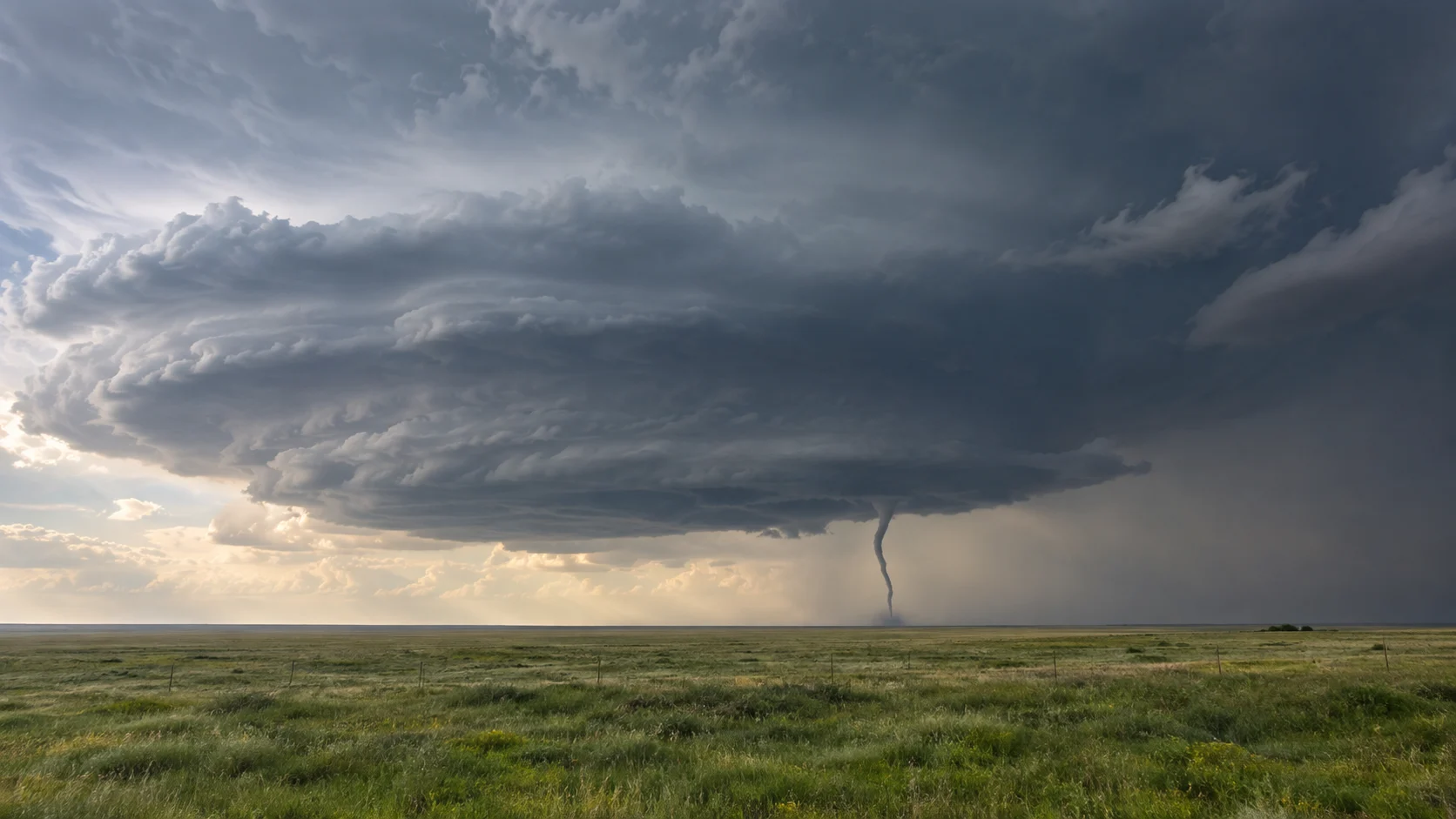 A slender tornado descends beneath a supercell over open prairie